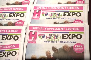Stack of pink-bordered flyers advertising a Senior Health Expo with 'FREE ADMISSION' circles and smiling people image across pages.