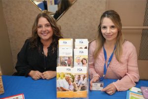Two women sit at a blue table with an FCA brochure display, smiling at the camera.