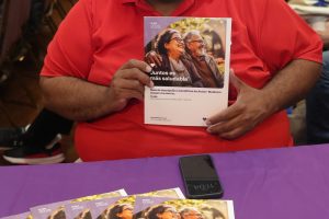 Smiling man in a red shirt sits at a table, holding a health brochure titled 'Juntos es más saludable' at a busy event with attendees in the background.