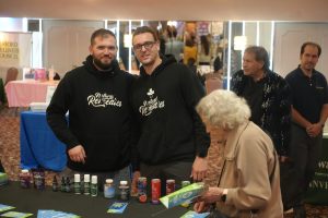Two men in matching black hoodies with 'Natural Remedies' stand behind a product table at a convention booth presenting bottles and cans of supplements.