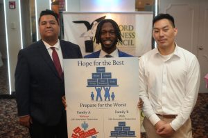 Three men in suits pose with a large life-insurance poster featuring a pyramid diagram in front of an event backdrop.