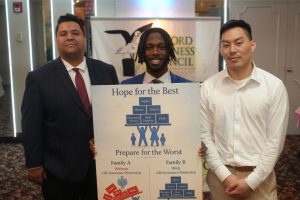 Three men in business attire pose with a poster that reads 'Hope for the Best ... Prepare for the Worst' at an event venue behind them.