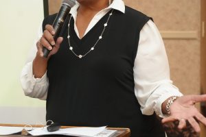 Woman speaking into a handheld microphone at a wooden podium, wearing a white blouse with a black vest and pearl necklace.