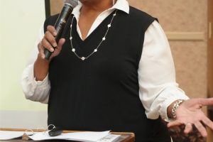 Woman speaking into a handheld microphone at a wooden podium during a presentation, wearing a white blouse and black vest with a pearl necklace.