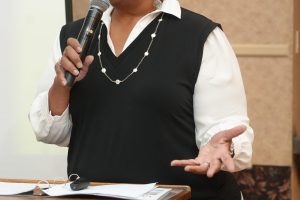 African American woman in a white blouse and black vest speaks into a handheld microphone at a wooden podium with papers in front.
