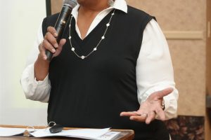 Woman speaking into a microphone at a podium during a presentation, gesturing with her hand.
