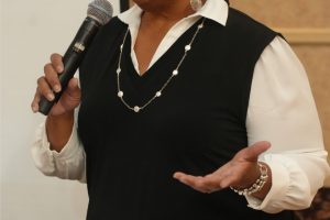 African American woman in a black vest over a white blouse speaks into a handheld microphone at a wooden podium with papers on its surface.