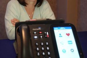 Smiling woman in a light sweater sits at a desk, with a large office phone in the foreground.