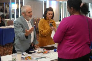Man in a light-blue blazer speaks and gestures to a woman in a mustard sweater at a conference booth; table has flyers and candies in front of them.