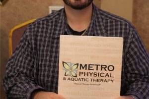 Man with glasses and beard sits at a blue table, holding a Metro Physical & Aquatic Therapy brochure toward the camera.