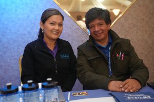 Two people sit at a table, smiling for the camera; woman in a black jacket with a MARK Medical Care logo beside a man with a pen-filled vest, blue backdrop behind them.