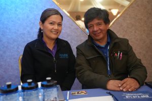 Smiling woman in a dark jacket sits beside a man with a blue lanyard at a booth table during an event.