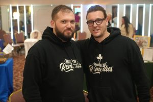 Two men in black hoodies pose together and smile for a photo at an indoor event space with bright vertical lights in the background.
