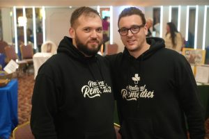 Two men in black hoodies with a 'Natural Remedies' logo pose together at an indoor event, smiling at the camera.