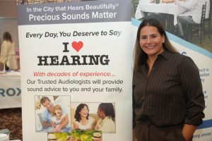 Smiling woman standing beside a banner that reads 'I love HEARING' at a hearing health event.
