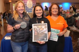 Three smiling women stand together at an event, holding brochures and a large printed page, with a crowd and blue table in the background.