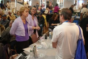 Two women in purple tops talk with a man in a white polo at a crowded expo booth.
