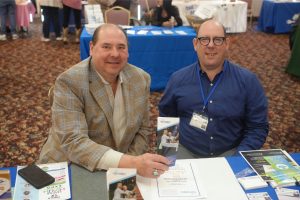 Two men at a conference table exchange a brochure; one wears a plaid beige blazer, the other a blue shirt with a name badge and glasses.