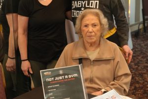 Three women posing indoors at a gym event: an elderly woman seated at a table flanked by two smiling younger women behind her.