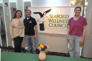 Three smiling adults stand indoors beside a Seaford Wellness Council banner, with a green table and colorful flowers in the foreground.