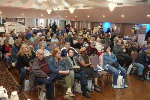 Audience seated in a large hall attending a community event or conference, with banners and booths in the background.”] ,