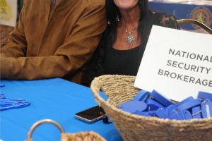 Two smiling adults sit at a blue-table booth; a sign reads 'National Security Brokerage' while baskets hold blue badges and white pins in front.