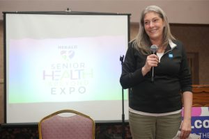 Smiling woman with a microphone speaks at a Senior Health & Beyond Expo presentation, with the event logo on the projector screen behind her.