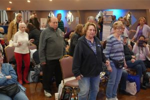 Group of older adults at an indoor community event, some standing among rows of chairs and bags on the floor.