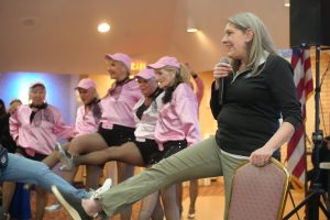 Woman with gray hair speaks into a microphone on stage, while a line of dancers in pink outfits and hats sit or stand behind her with smiles, an American flag visible in the background.