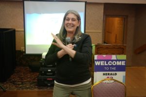 Smiling woman holds a microphone and speaks at a conference, with a projector screen behind her and a welcome sign nearby.