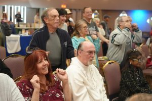 A crowded indoor event with people seated and standing, smiling and watching something off-camera.
