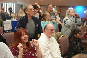 A cheerful group of people seated and standing at a social event in a hall, smiling and watching something off-camera.