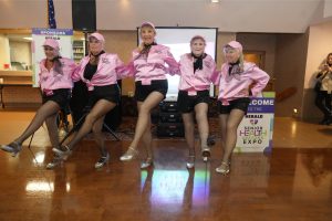 Five women in pink satin jackets perform a synchronized kickline on a stage at a senior health expo, with a projector screen behind them.
