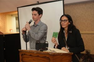 Two presenters at a wooden podium during a formal event; a man speaks into a handheld microphone while a woman beside him shows a green card or badge.