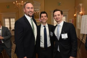 Three men in dark suits smile for a group photo at an indoor formal event, chandeliers overhead and warm lighting behind them.