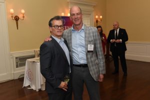 Two men in business attire pose for a photo at a reception; one holds a wine glass and wears a name badge, with another attendee in the background.