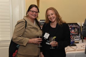 Two smiling women at a social event pose for a photo, each holding a glass of beverage; one in a beige blazer with glasses, the other in a black blazer with a name badge holding a lime-edged drink.