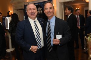 Two men in suits posing and smiling at a formal event, one holding a beer bottle and wearing a name badge in a busy backdrop.