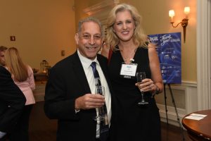 Two adults in formal attire pose together at a reception, each holding a champagne flute and smiling.
