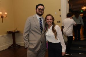 Two people posing for a photo at an indoor event: a man in a light gray suit and purple striped tie next to a smiling woman in a white blouse and dark pants.