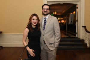 Couple posing together at a formal event: woman in a black dress and red lipstick, man in a light gray suit with a striped tie, smiling in an elegant hallway.