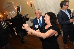 Woman in a black dress holds up a large black case at a formal reception, smiling as guests chat in the background.