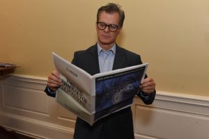 Man in a dark suit and glasses reads a newspaper in a beige hallway.