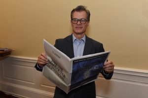 Man in a suit and glasses reads a newspaper indoors, smiling slightly at the camera.