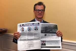 Man in a dark suit and glasses holding an open newspaper, showing a 'Top Lawyers' awards page on one side and a large article page on the other.