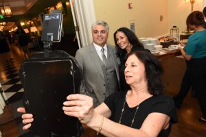 Woman in a black top holds a large black photo booth device at a buffet-style party, posing for a snap with guests nearby.