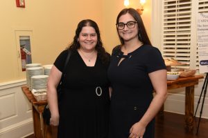 Two women in black dresses smiling for a photo at a catered event, standing near a buffet table with plates in the background.