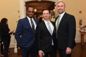 Three men in suits posing for a photo at an indoor reception, smiles for the camera in a doorway.