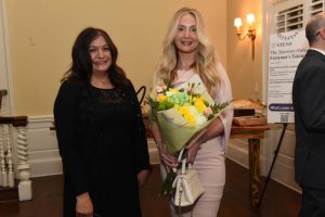 Two smiling women at an indoor event; the blonde woman in a pale pink dress holds a bouquet of yellow and white flowers.