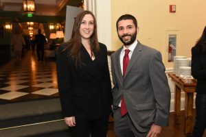 Two well-dressed individuals posing and smiling at a formal event in a lobby area with checkered flooring and a serving table in the background.